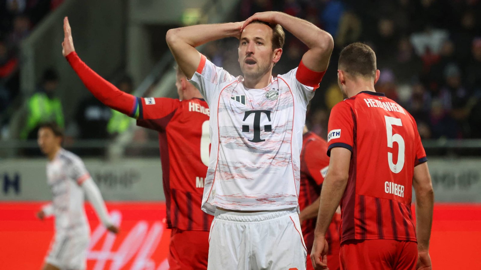Harry Kane du Bayern Munich semble frustré après un match, portant un maillot blanc avec des accents rouges, au milieu des joueurs de l'équipe adverse.