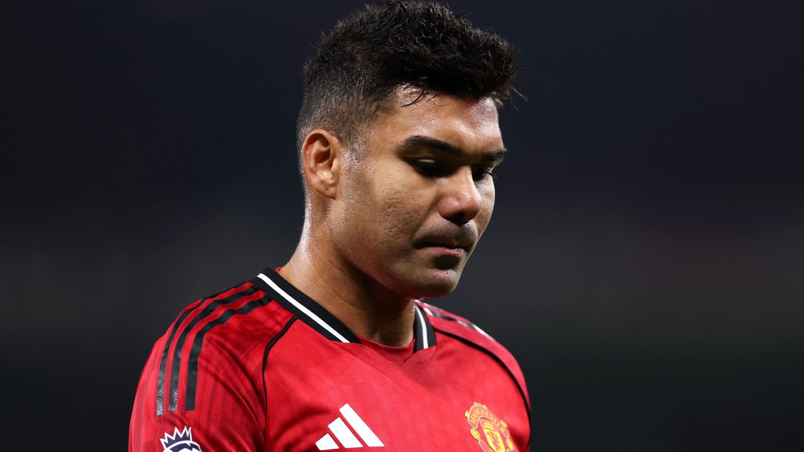 Casemiro, in een rood Manchester United-tenue, kijkt peinzend naar beneden, wat intensiteit weerspiegelt tijdens een wedstrijd tegen een donkere achtergrond.
