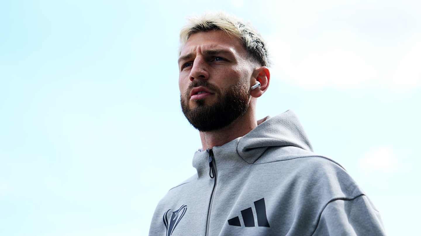 Un joueur de football confiant, portant un sweat à capuche gris et des écouteurs, regardant vers le haut contre un ciel bleu clair, affichant une expression concentrée.