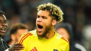 Excited football player in a yellow kit with red accents, celebrating passionately with a backdrop of blurred teammates in action.