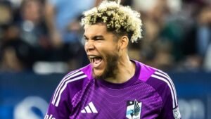 Feestend in een paars keepers tenue, juicht een speler met krullend blond haar gepassioneerd, wat de opwinding van een wedstrijdmoment belichaamt.