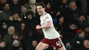 Declan Rice juicht vreugdevol na het scoren van een doelpunt, gekleed in een wit Arsenal-tenue met kastanjebruine korte broek, te midden van een juichende menigte.