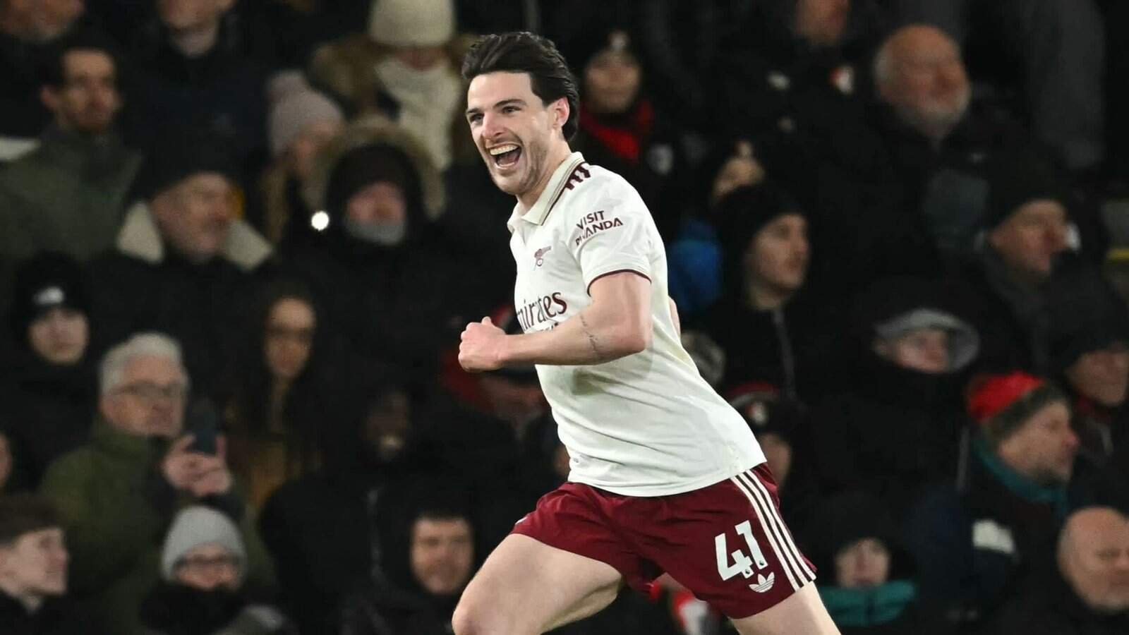 Declan Rice juicht vreugdevol na het scoren van een doelpunt, gekleed in een wit Arsenal-tenue met kastanjebruine korte broek, te midden van een juichende menigte.
