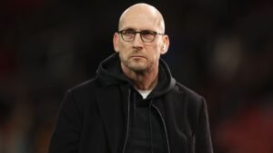 Jaap Stam in een zwarte jas en hoodie, met een serieuze uitdrukking, vastgelegd tijdens een voetbalwedstrijd.