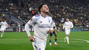 Jadon Sancho viert een doelpunt voor Aston Villa in een Europese wedstrijd, gekleed in een wit tenue met blauwe accenten, tegen een levendige menigte als achtergrond.