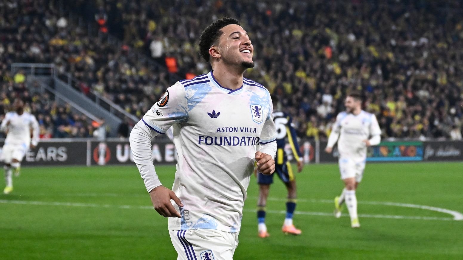 Jadon Sancho viert een doelpunt voor Aston Villa in een Europese wedstrijd, gekleed in een wit tenue met blauwe accenten, tegen een levendige menigte als achtergrond.