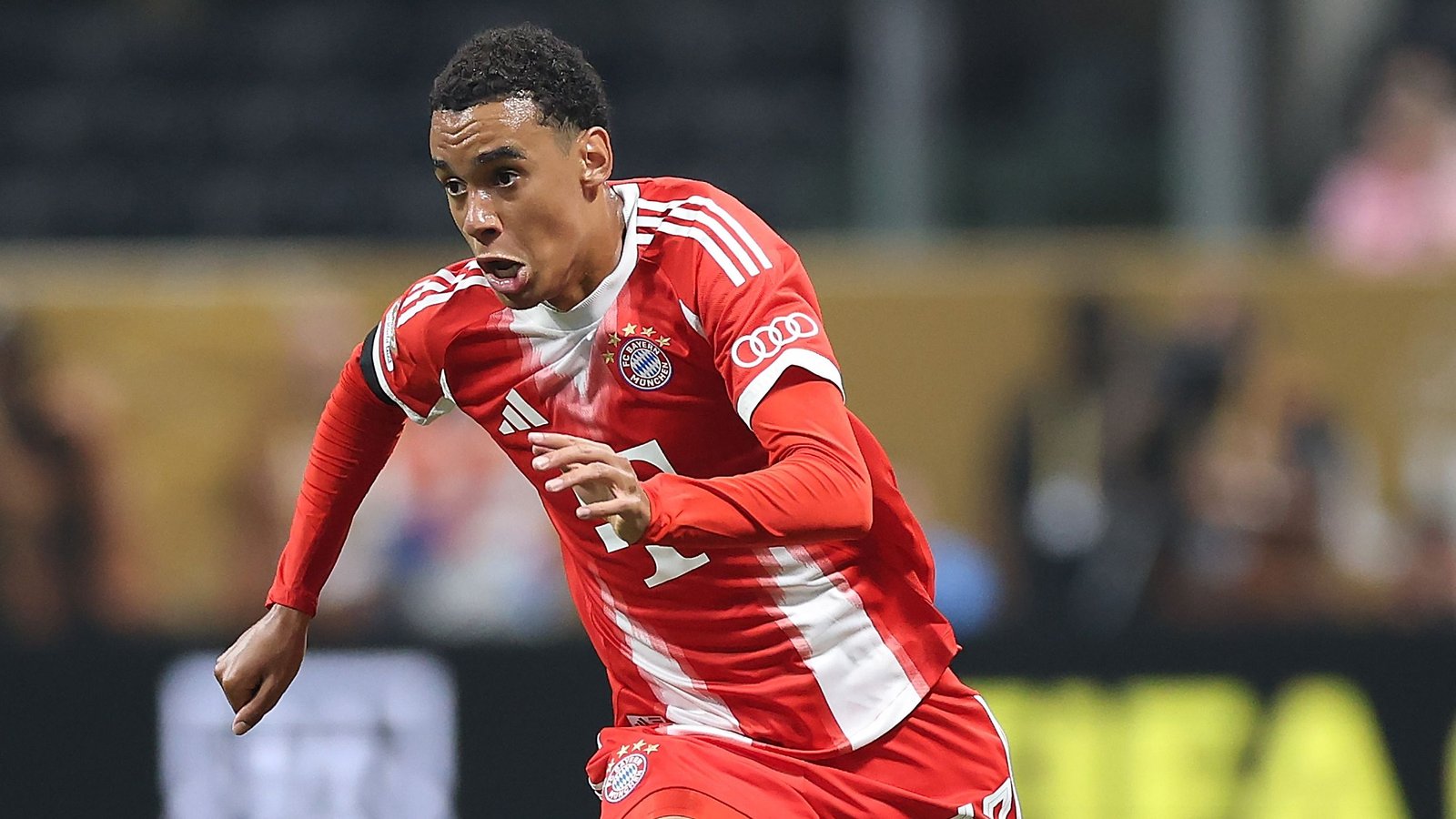 Jamal Musiala sprint vastberaden in het kenmerkende rood-witte tenue van Bayern München tijdens een intense wedstrijdactie.