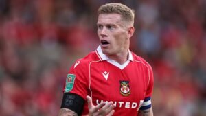 James McClean gesturing during a match for Wrexham, wearing a red kit with the club badge and captain's armband, against a blurred crowd background.