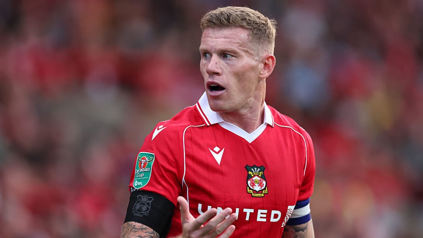 James McClean gesturing during a match for Wrexham, wearing a red kit with the club badge and captain's armband, against a blurred crowd background.