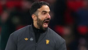 Ruben Amorim die gepassioneerd staat te schreeuwen aan de zijlijn, gekleed in een grijze jas met een leeuwembleem, tijdens een gespannen wedstrijdsfeer.