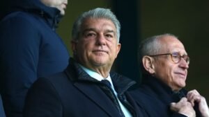 Joan Laporta, in een donkere jas, kijkt aandachtig vanaf de tribune, naast een andere man, tegen de achtergrond van een voetbalstadion.