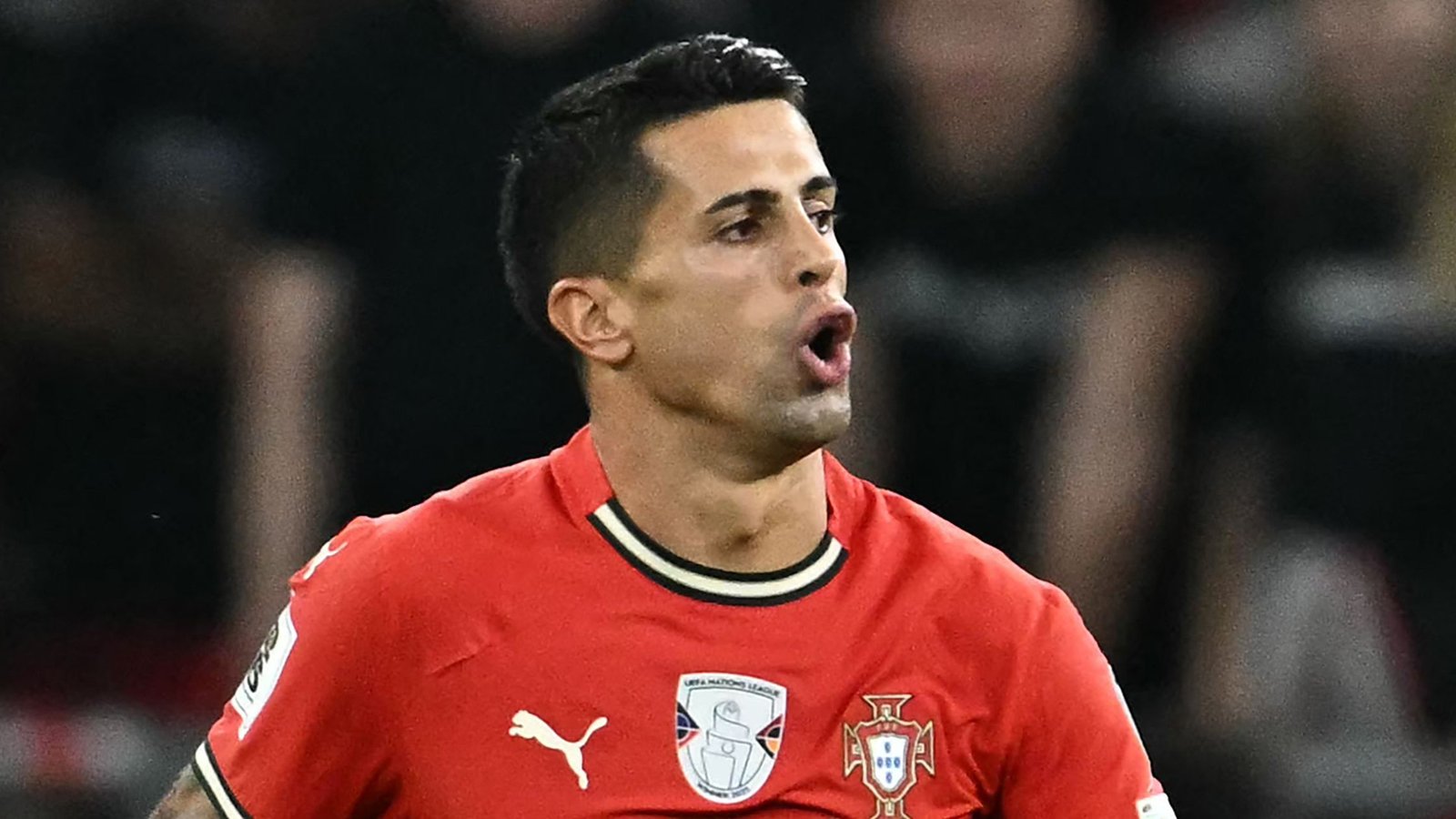 João Cancelo in een rood Portugal-tenue, gepassioneerd reagerend tijdens een UEFA Nations League-wedstrijd, met een geconcentreerde gezichtsuitdrukking.