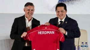 John Herdman glimlacht terwijl hij een rood tenue vasthoudt met "HERDMAN" erop gedrukt, naast een official in een donker pak tijdens een persevenement.