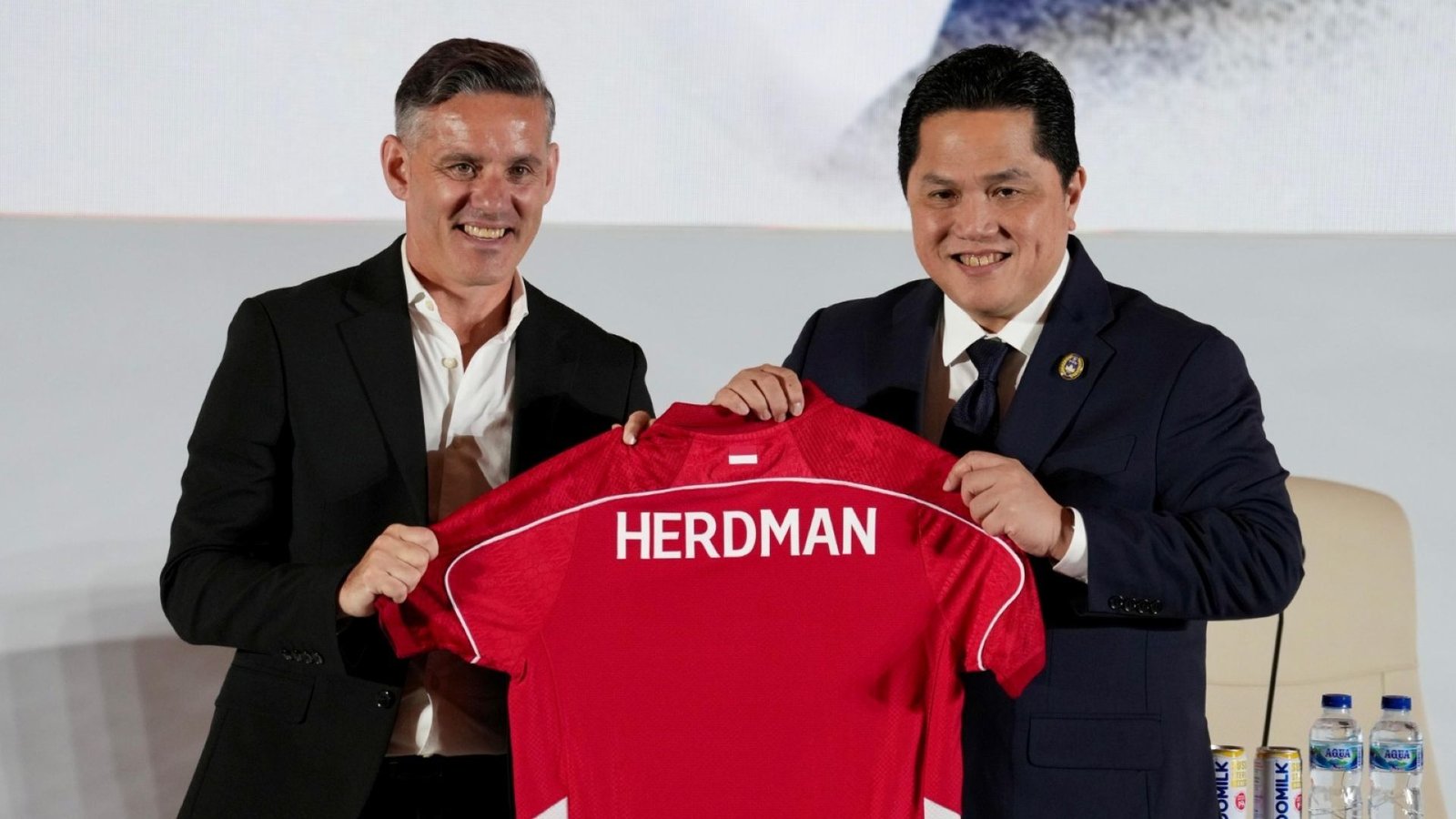 John Herdman glimlacht terwijl hij een rood tenue vasthoudt met "HERDMAN" erop gedrukt, naast een official in een donker pak tijdens een persevenement.
