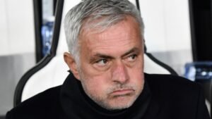 Jose Mourinho ziet er gespannen uit met een ernstige uitdrukking, gezeten in een donker jasje tegen een wazig stadionachtergrond.