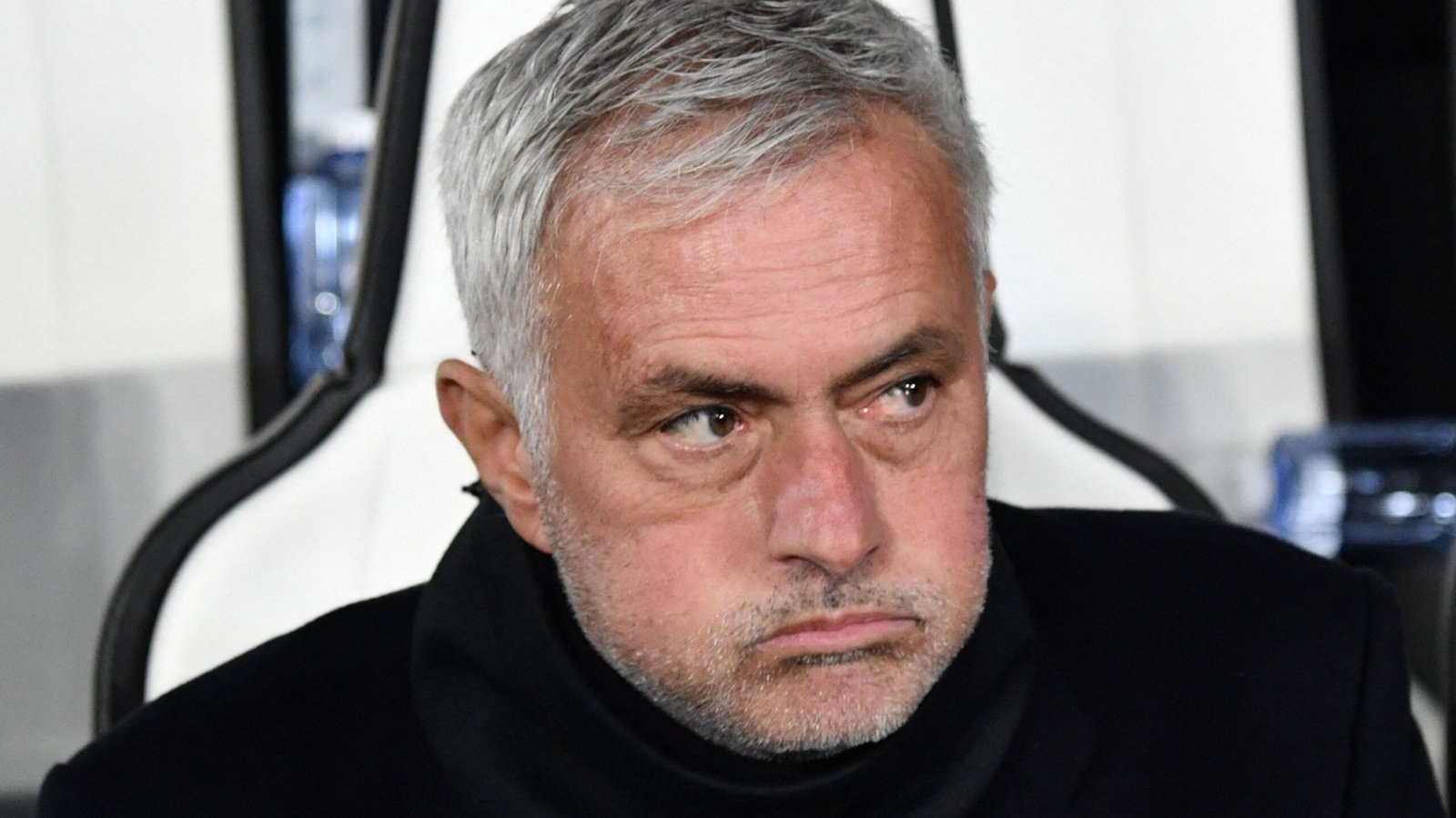 Jose Mourinho ziet er gespannen uit met een ernstige uitdrukking, gezeten in een donker jasje tegen een wazig stadionachtergrond.