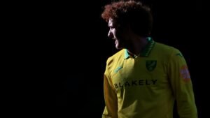 Josh Sargent in een felgeel Norwich City-tenue, met de rug naar de camera, tegen een donkere achtergrond.