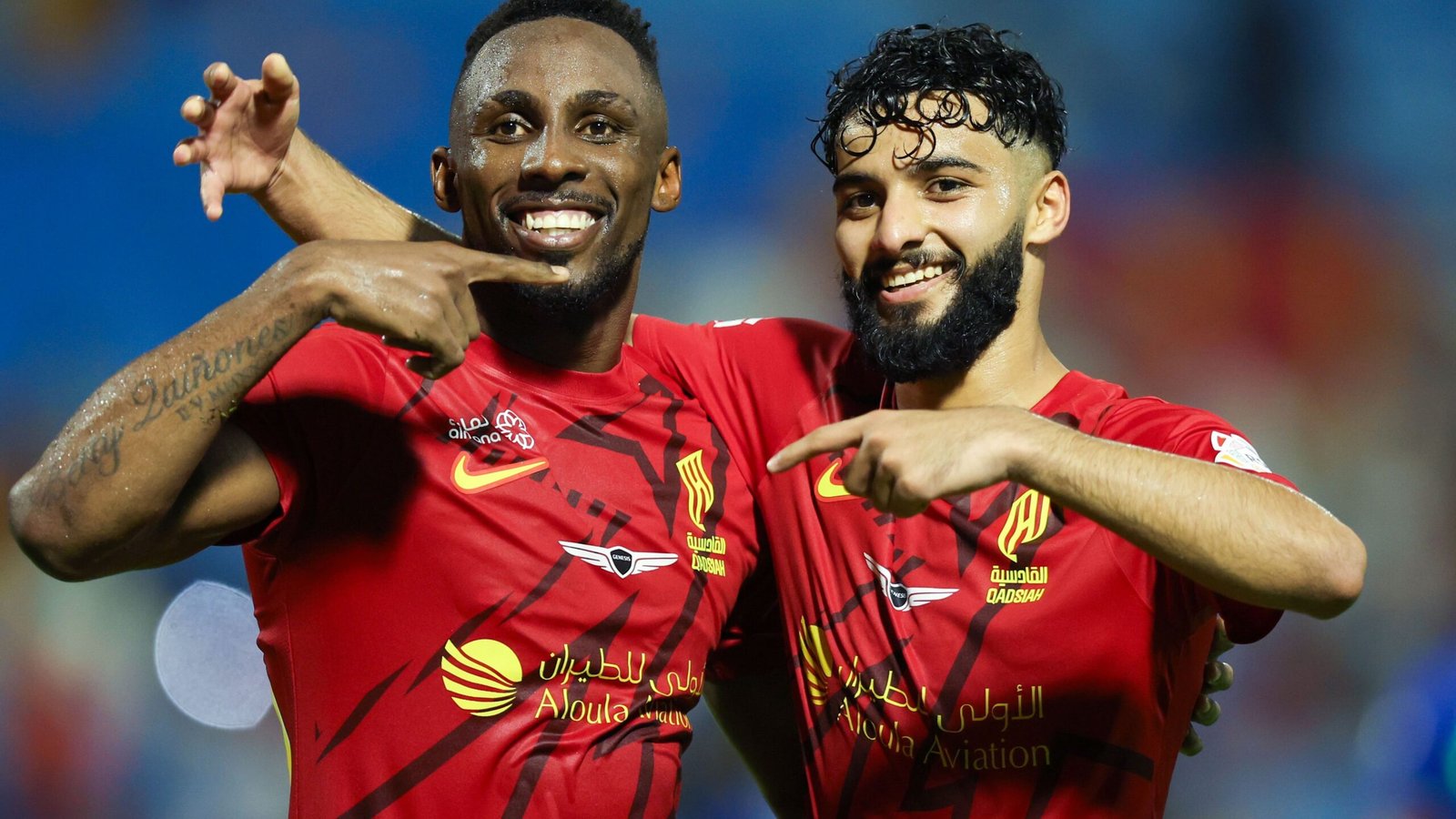 Julian Quinones en een teamgenoot vieren een doelpunt, wijzen naar elkaar, gekleed in het rode thuistenue van Al-Wehda in een levendig stadion.