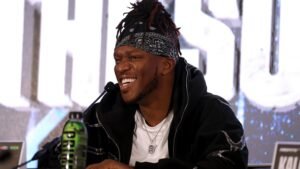 KSI glimlacht tijdens een persconferentie, draagt een zwarte bandana en hoodie, met een groene drinkfles zichtbaar naast hem.