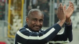 Rhulani Mokwena sourit et applaudit, vêtu d'un pull à rayures bleu marine et blanc, au milieu d'une foule enthousiaste lors d'un match de football.