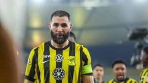 Karim Benzema in zijn zwarte en gele Ittihad Club-outfit, gefocust tijdens een wedstrijd, met teamgenoten zichtbaar op de achtergrond.