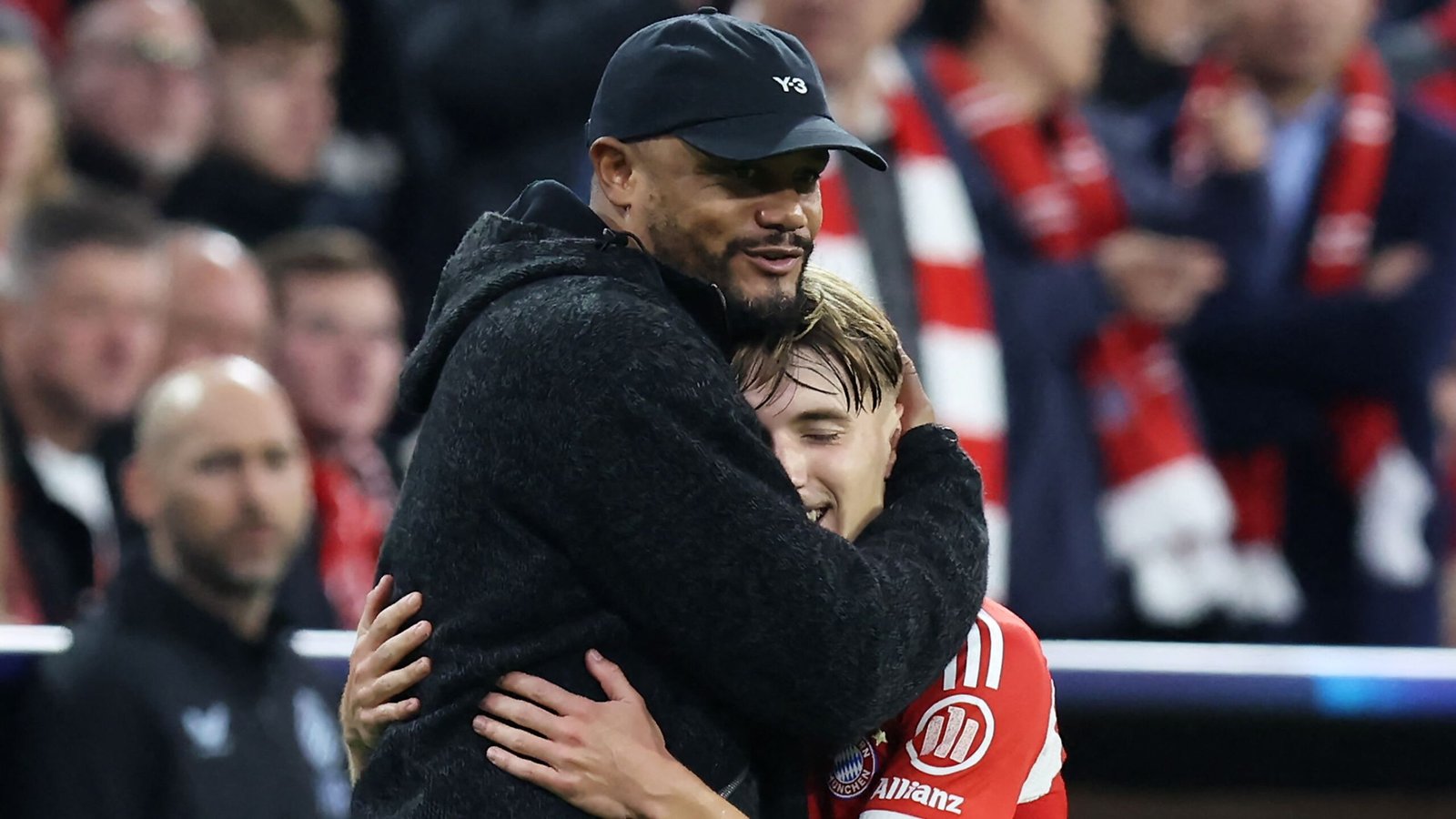 Kompany omhelst een lachende jonge speler in een Bayern Munich-tenue, met fans op de achtergrond, terwijl ze een wedstrijdzege vieren.