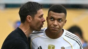 Kylian Mbappé kijkt aandachtig toe terwijl Xabi Alonso dichtbij hem spreekt, beiden in voetbalkleding, tegen een gele achtergrond van een wedstrijd.