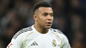 Kylian Mbappé portant le maillot blanc du Real Madrid, l'expression concentrée pendant un match, avec les lumières du stade floues en arrière-plan.