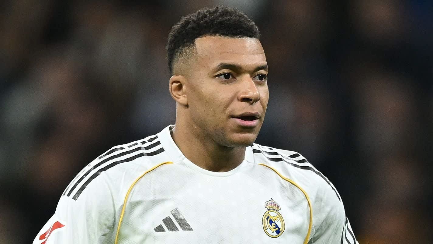 Kylian Mbappé portant le maillot blanc du Real Madrid, l'expression concentrée pendant un match, avec les lumières du stade floues en arrière-plan.