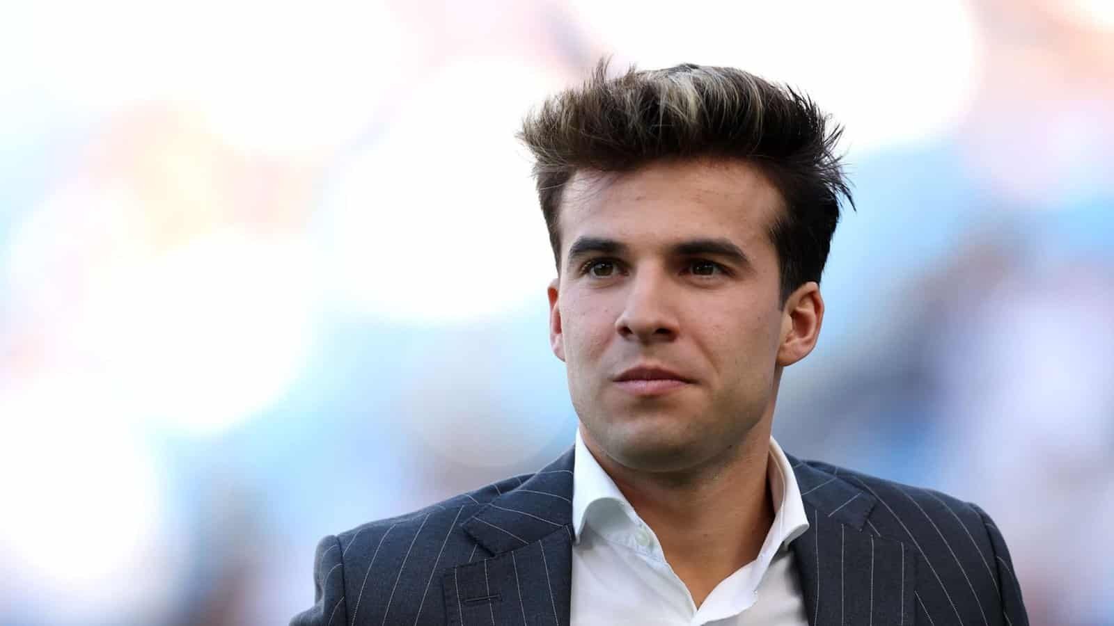 Riqui Puig, en costume rayé sombre, regarde pensivement devant lui avec un arrière-plan de stade flou, réfléchissant à son revers lié à l'opération du LCA.