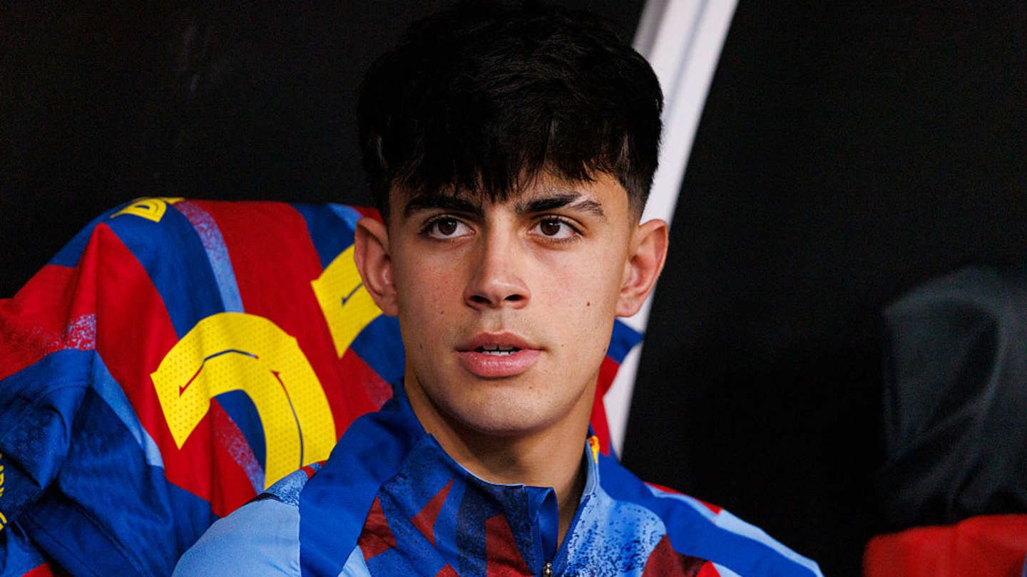 Young footballer seated on the bench, wearing a blue and red FC Barcelona tracksuit, with a focused expression against a dark background.
