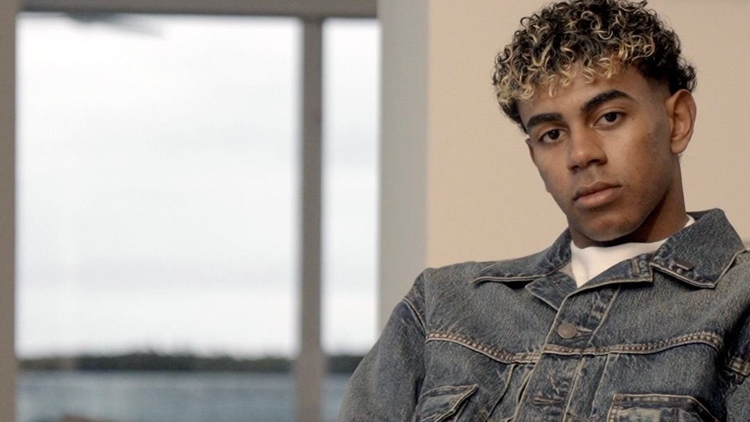 Lamine Yamal, wearing a denim jacket, gazes thoughtfully at the camera, set against a softly lit indoor background.