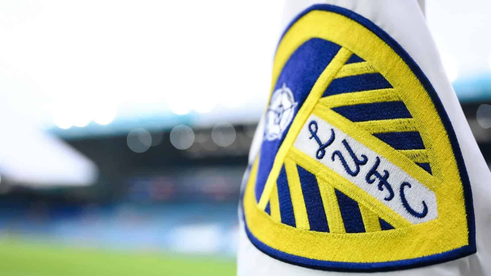 Het embleem van Leeds United is prominent weergegeven in blauw en geel, met een gestileerd ontwerp met ingewikkelde details.