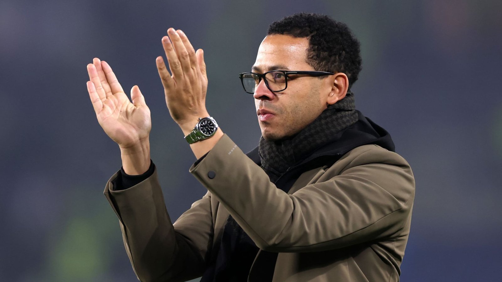 ليام روزينيو، يرتدي معطفاً بنياً ووشاحاً أسود، يصفق بحماس معبراً عن التشجيع خلال مباراة.