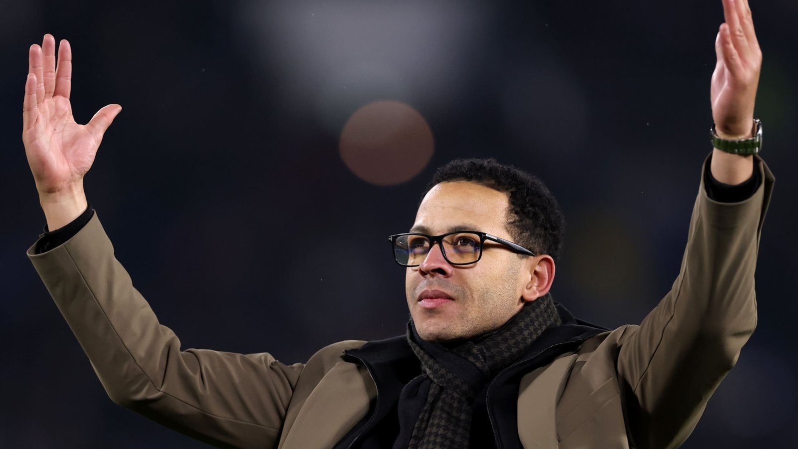 Liam Rosenior salue les supporters, les bras levés, vêtu d'un manteau marron et d'une écharpe noire, devant l'arrière-plan flou d'un stade.