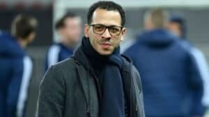 Liam Rosenior, portant des lunettes et une écharpe sombre, se tient dans un cadre de football, avec des coéquipiers flous en arrière-plan.