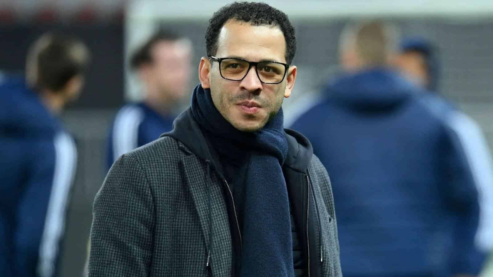 Liam Rosenior, portant des lunettes et une écharpe sombre, se tient dans un cadre de football, avec des coéquipiers flous en arrière-plan.