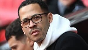 Liam Rosenior kijkt bedachtzaam, met een bril en een witte hoodie onder een zwarte jas, tegen de achtergrond van een voetbalstadion.