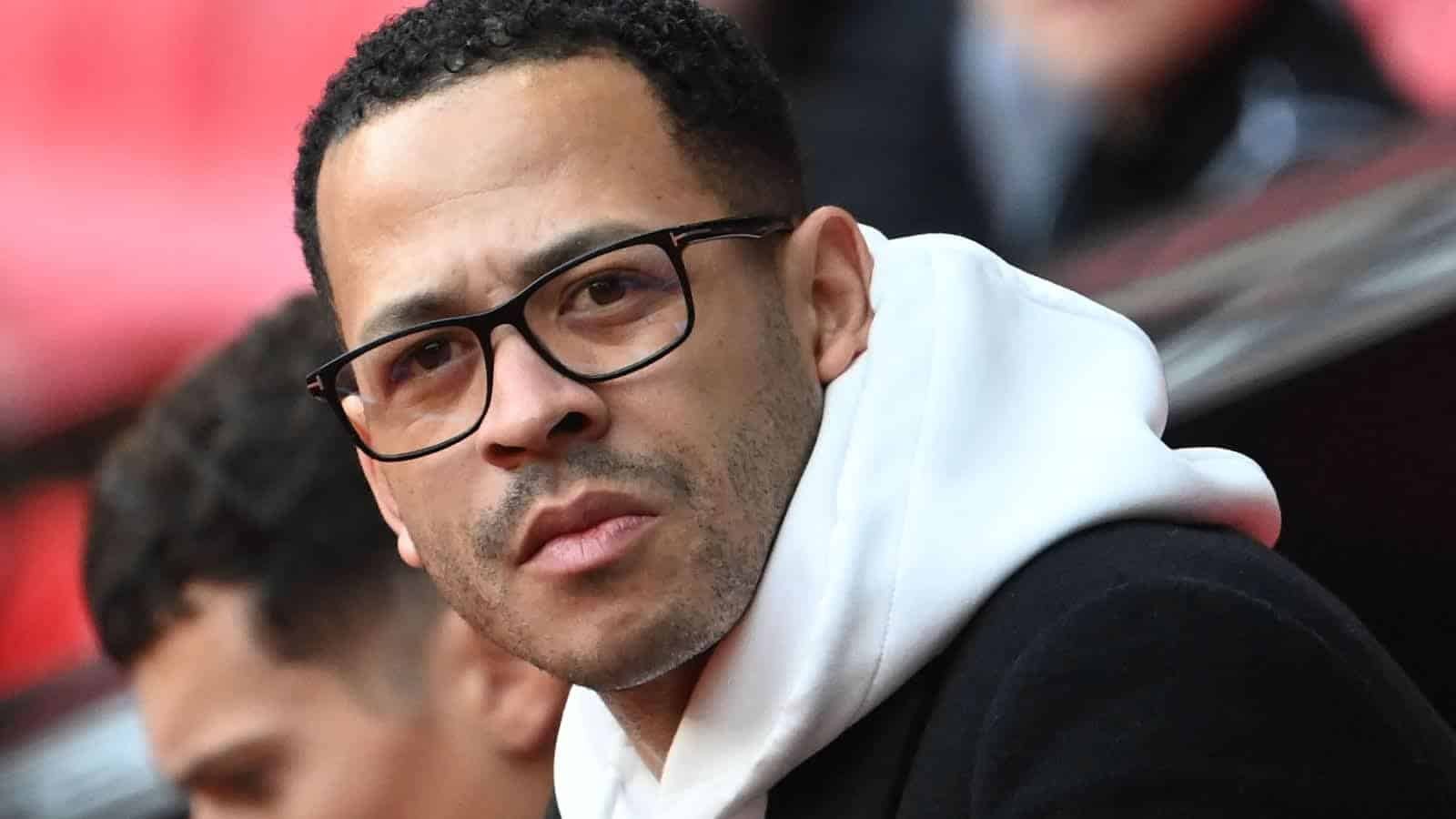 Liam Rosenior kijkt bedachtzaam, met een bril en een witte hoodie onder een zwarte jas, tegen de achtergrond van een voetbalstadion.