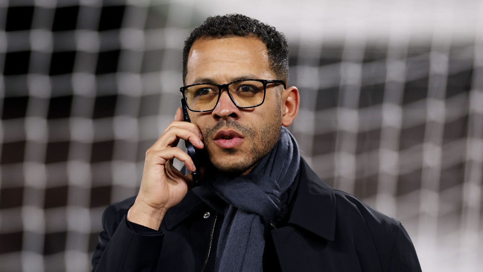 Liam Rosenior spreekt aan de telefoon, draagt een bril en een donkere jas, voor een voetbaldoelnet.