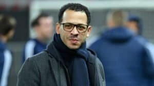 Liam Rosenior, met een bril en een donkere sjaal, glimlacht zelfverzekerd tegen een wazige voetbaltrainingsachtergrond.