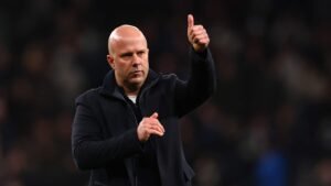 Entraîneur en manteau sombre faisant un geste de pouce levé, montrant son encouragement devant un fond de stade flou, lors d'un moment d'après-match.