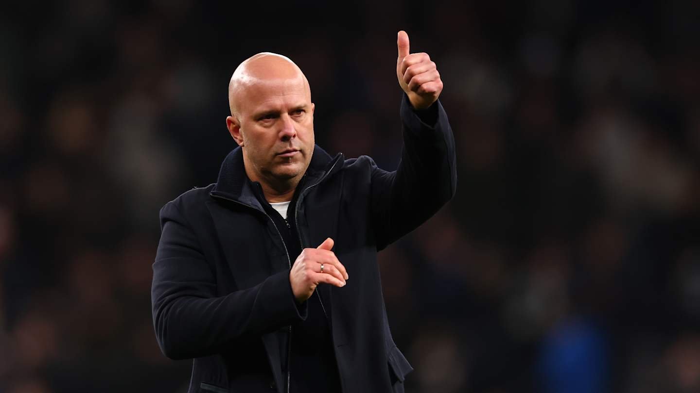 Entraîneur en manteau sombre faisant un geste de pouce levé, montrant son encouragement devant un fond de stade flou, lors d'un moment d'après-match.