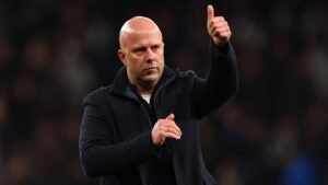 Entraîneur faisant un geste de pouce levé dans une veste sombre, avec une expression concentrée, au milieu d'un arrière-plan flou de foule dans le stade.