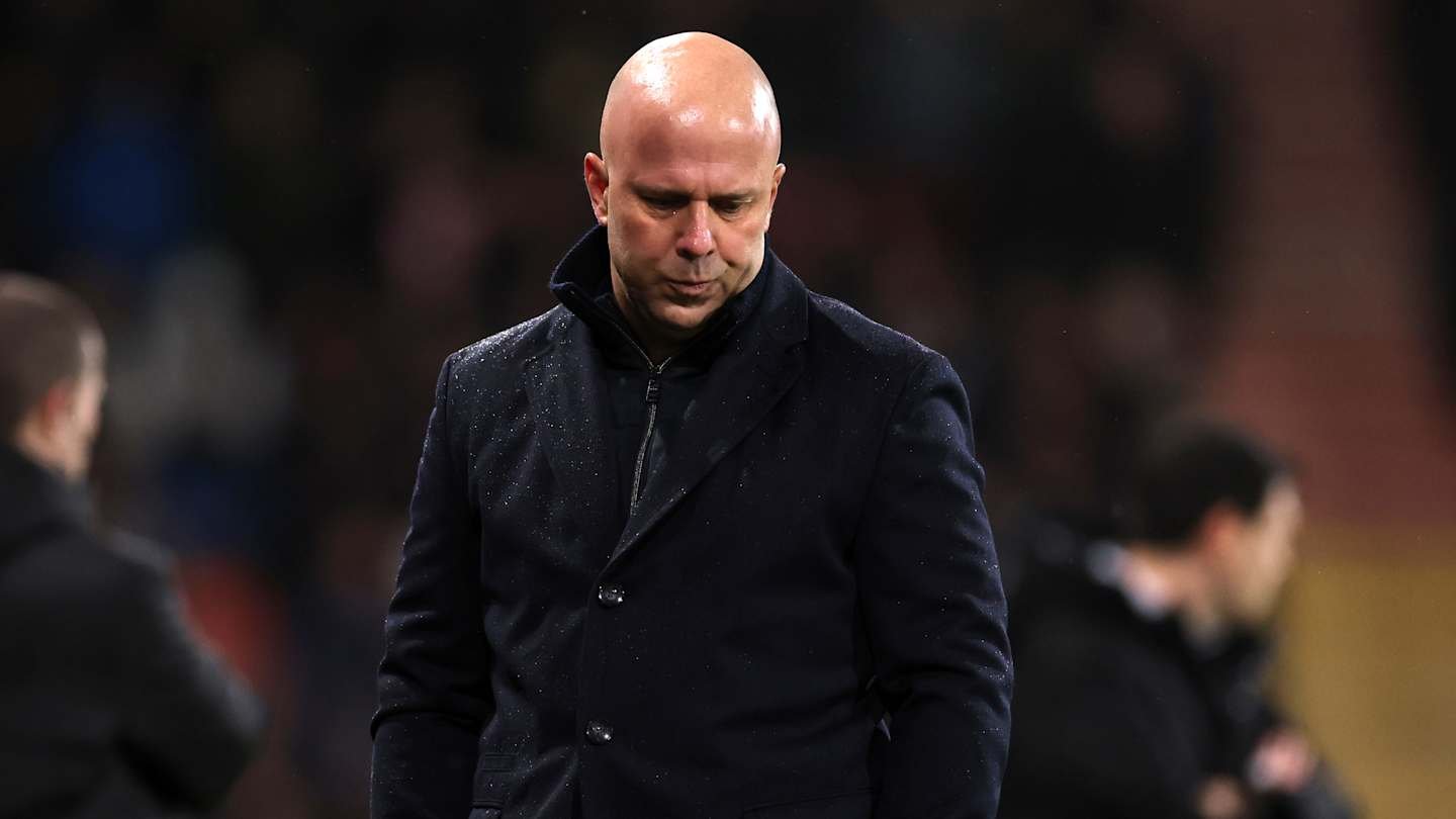 Liverpool manager looks dejected on the sidelines, wearing a dark coat, amid a tense atmosphere during a pivotal match.