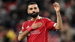 Mohamed Salah, in a red Liverpool kit, raises his hand while gesturing to fans, showcasing his signature beard and focused expression.