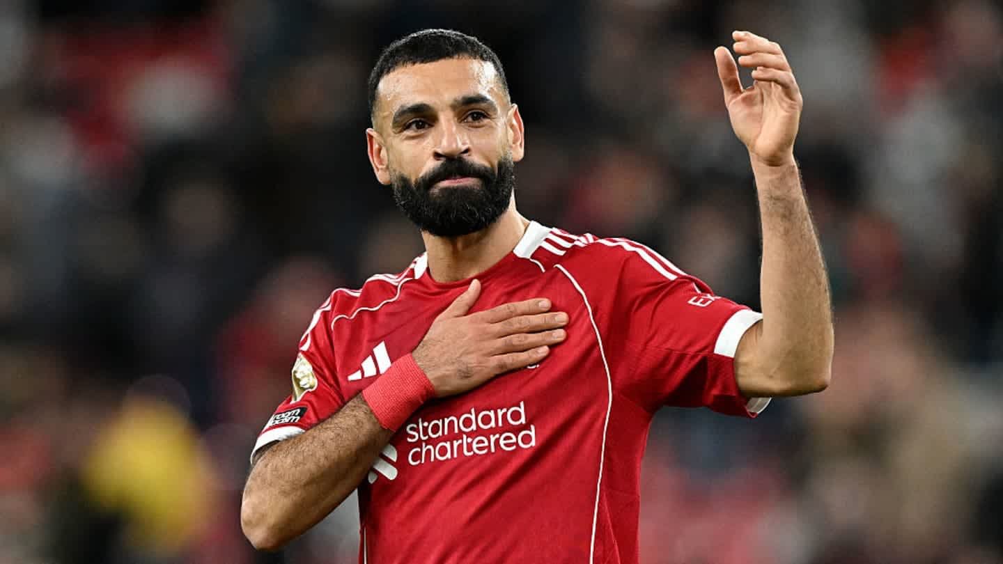 Mohamed Salah, in a red Liverpool kit, raises his hand while gesturing to fans, showcasing his signature beard and focused expression.