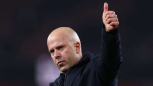 Manager giving a thumbs-up gesture, wearing a dark jacket, with a stadium backdrop, conveying confidence and approval.