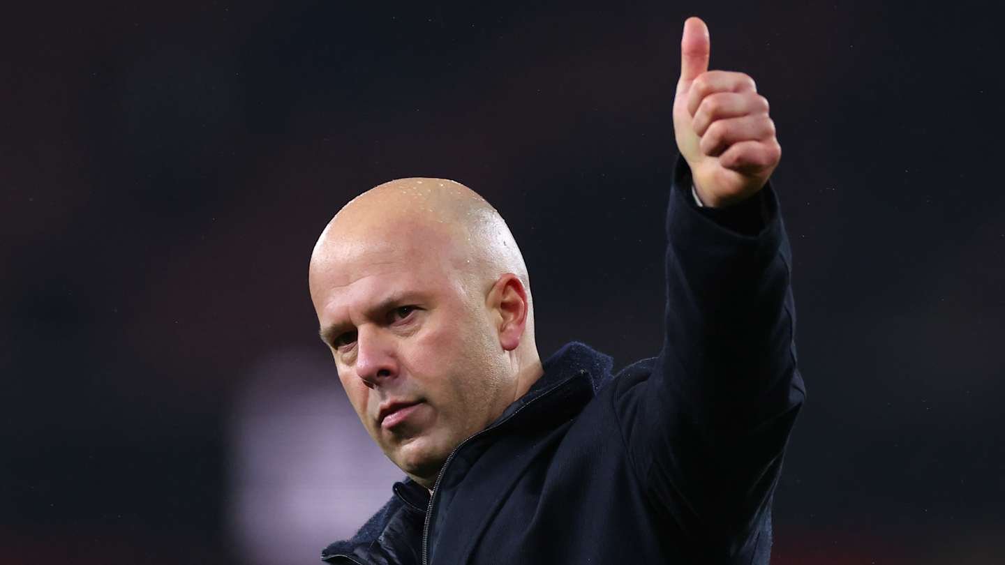 المدير يؤدي إيماءة بالإبهام للأعلى، مرتدياً سترة داكنة، مع خلفية ملعب، لنقل الثقة والموافقة.