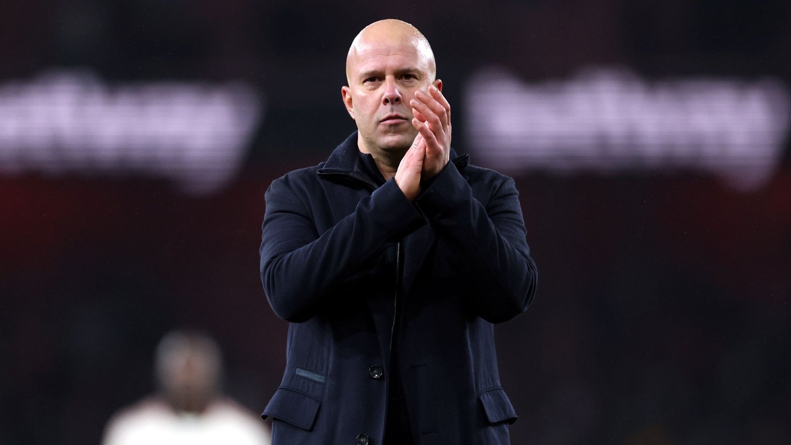 Coach applauding on the touchline, wearing a dark coat, focused expression, set against a blurred stadium backdrop during a match.
